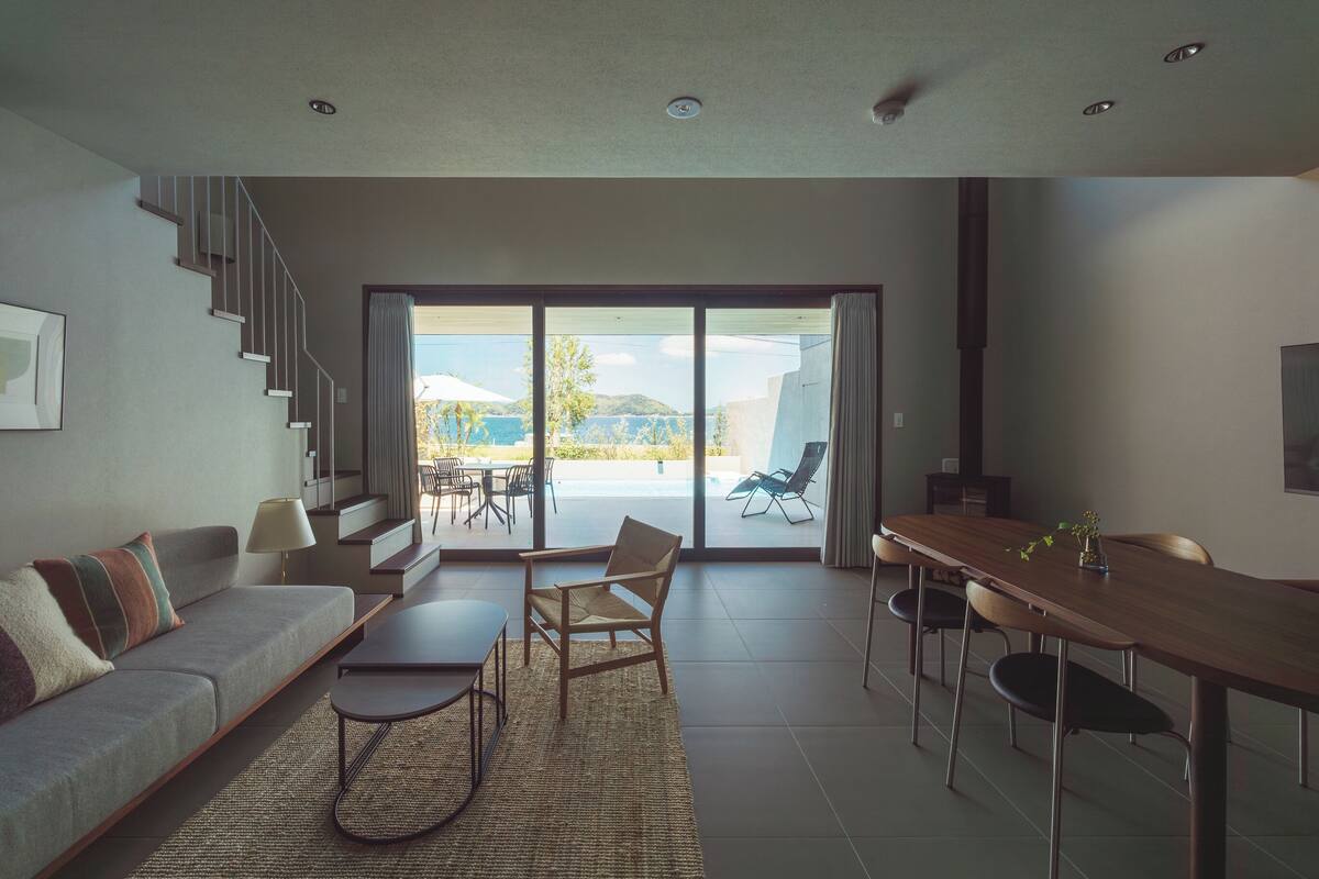 Una sala de estar moderna con un sofá, una mesa de centro y una mesa de comedor. Una escalera conduce a un balcón con vista al mar.