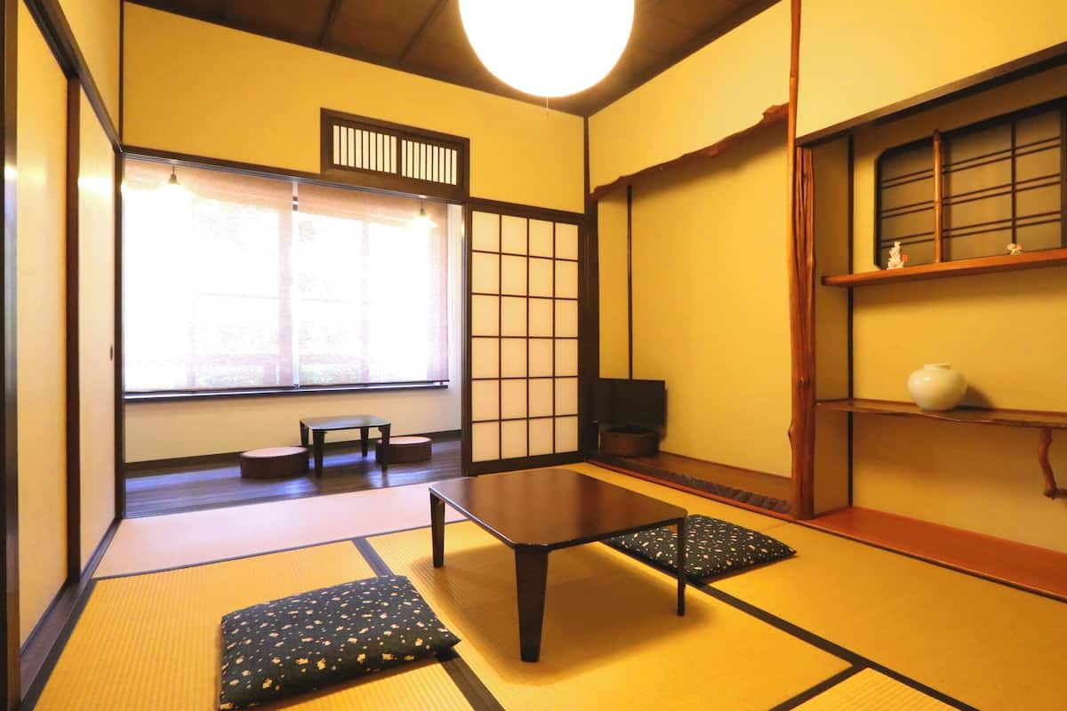 A tatami mat room with a low table, cushions, a sliding door, and a shelf with a vase.