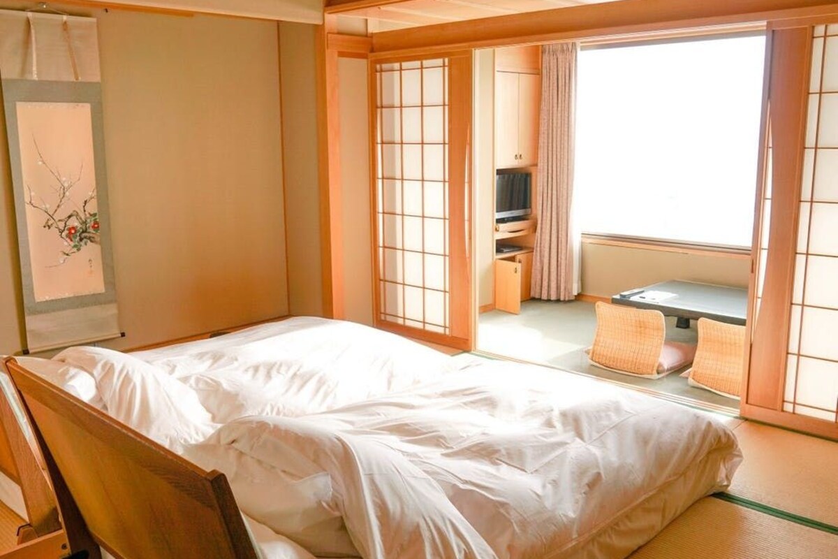 A traditional Japanese room with tatami flooring, a futon bed, sliding shoji doors, and a view of a garden.