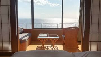A room with a bed, a wicker sofa, a small table, and a view of the sea through a large window.