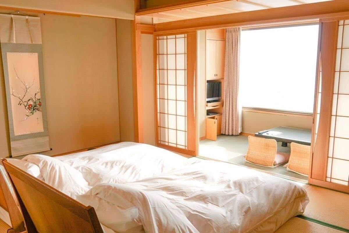 A traditional Japanese room with tatami flooring, a futon bed, sliding shoji doors, and a view of a garden.
