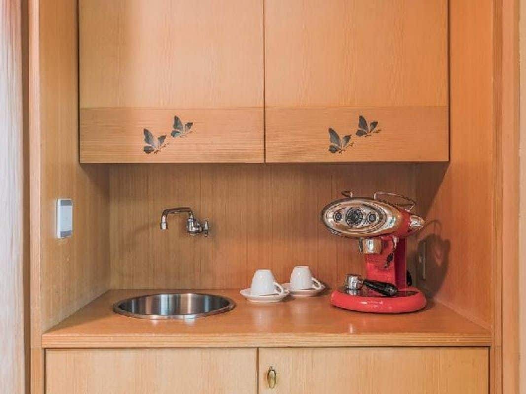 Une petite cuisine équipée avec un évier en acier inoxydable, une cafetière rouge et deux tasses blanches posées sur un plan de travail en bois.