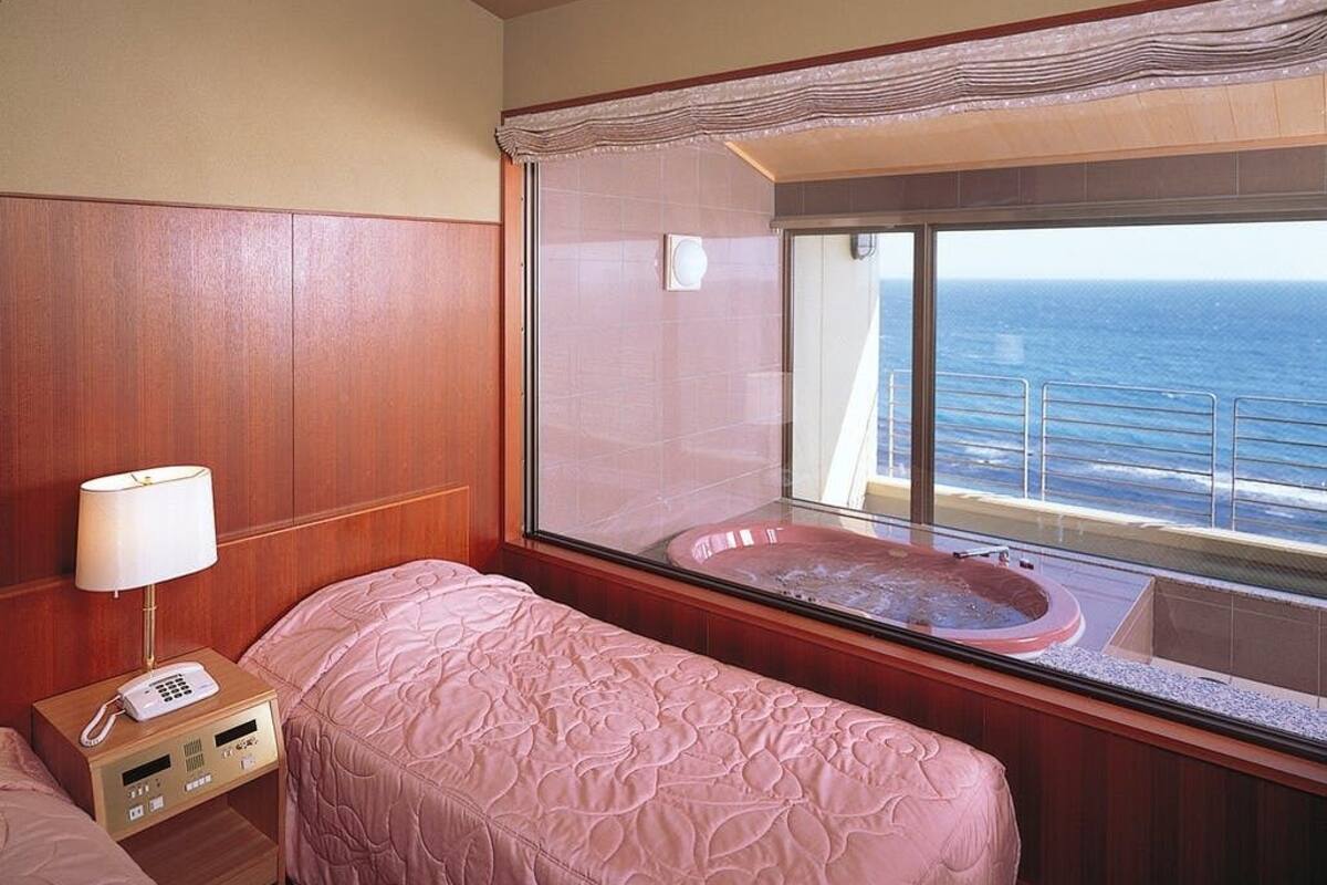 A room with a bed, a telephone, a window with a view of the ocean, and a hot tub.