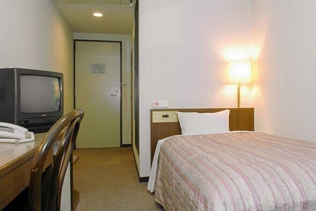 A hotel room with a bed, a desk with a television, and a nightstand with a lamp.