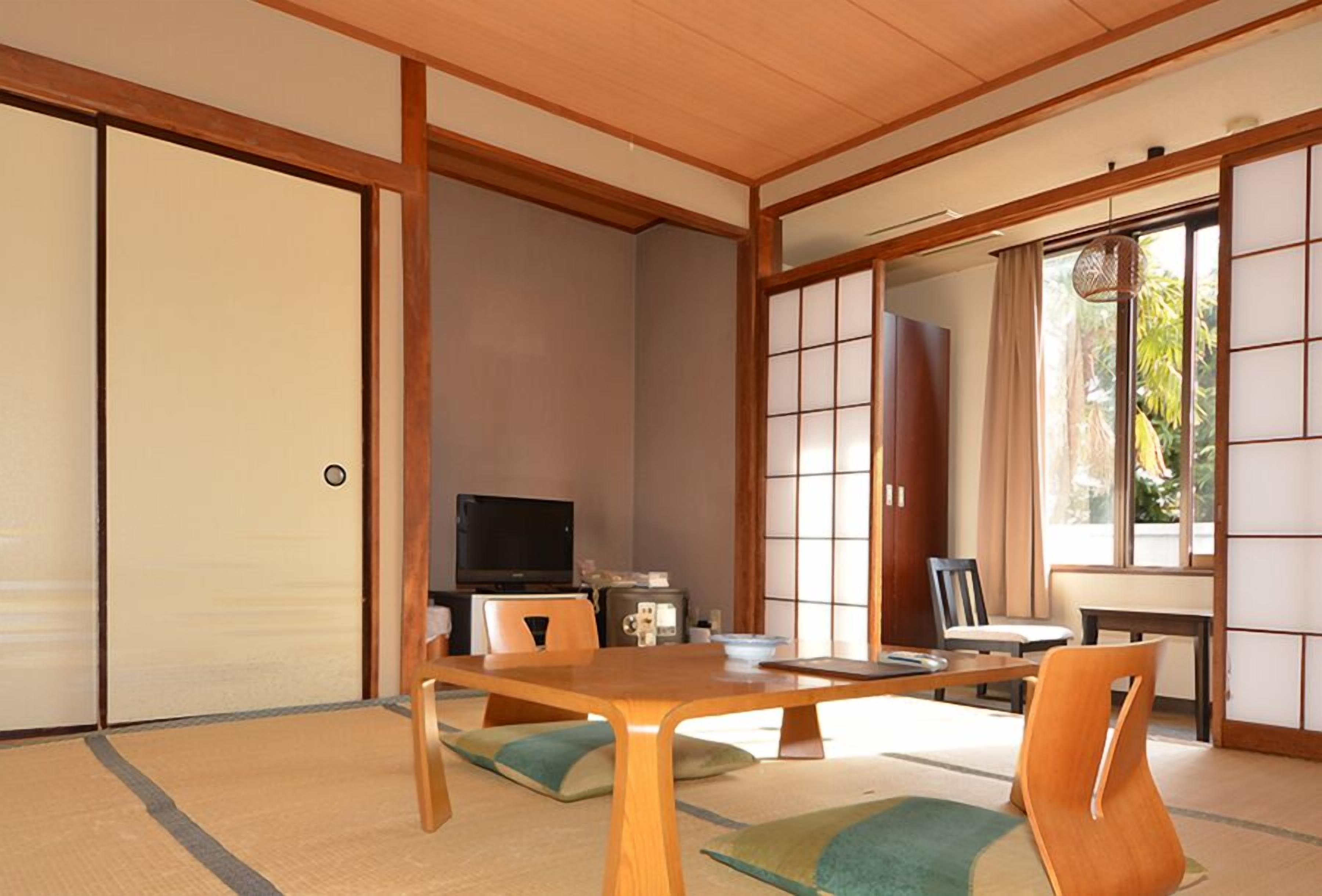 A traditional Japanese room with tatami flooring, sliding doors, a television, and a dining table with chairs.