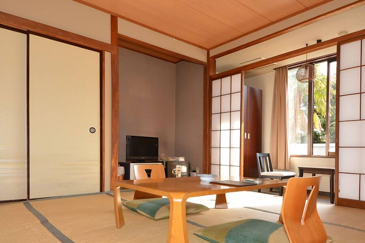 A traditional Japanese room with tatami flooring, sliding doors, a television, and a dining table with chairs.