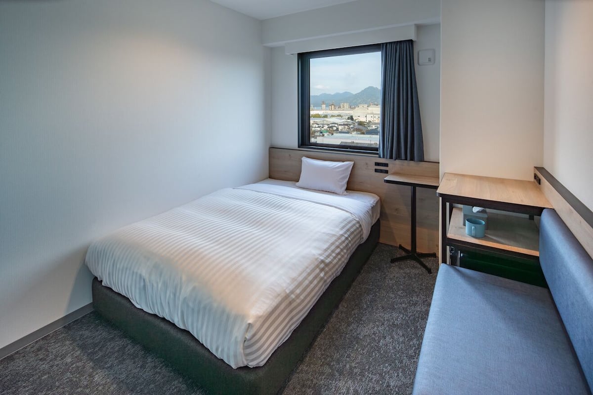 A hotel room with a bed, a small desk, a chair, a window with a view, and a carpeted floor.