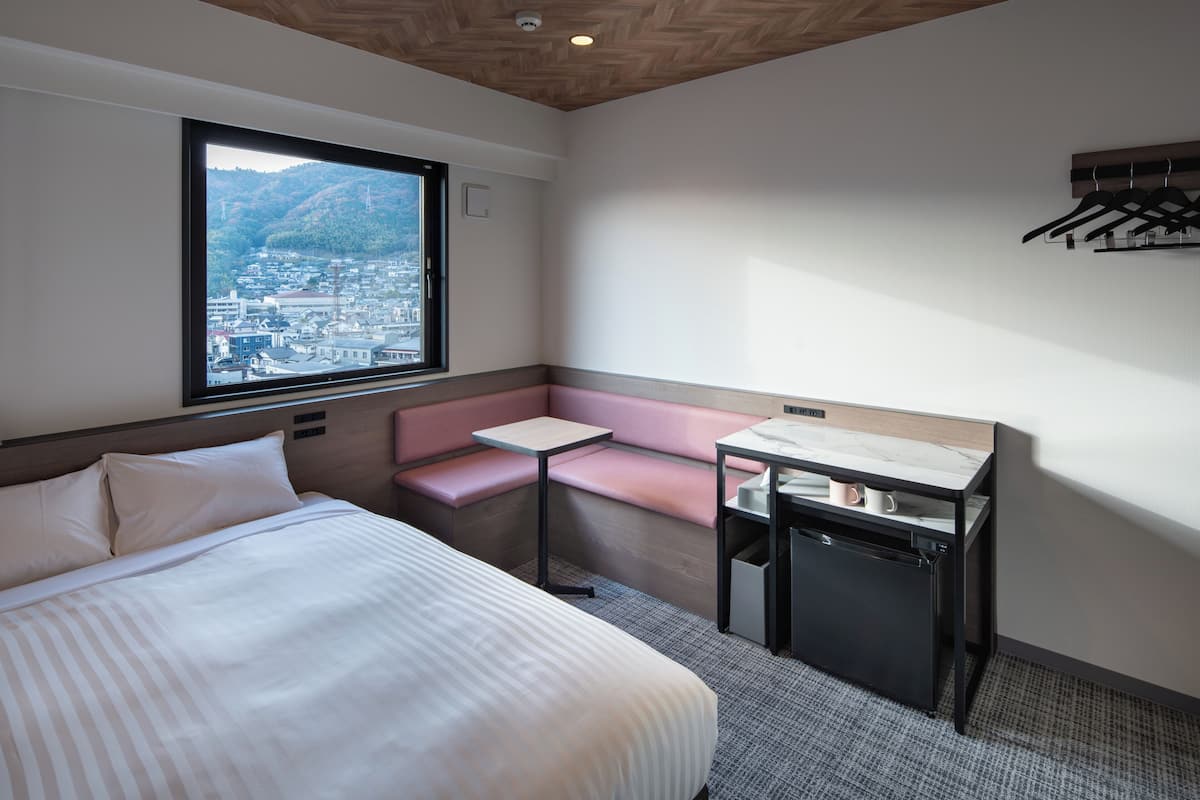 A hotel room with a large bed, a small table, a bench, a window with a city view, and a coat rack.