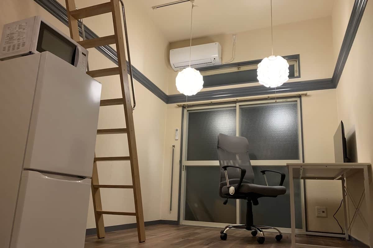 A room with a refrigerator, a chair, a ladder, an air conditioner, and a window.