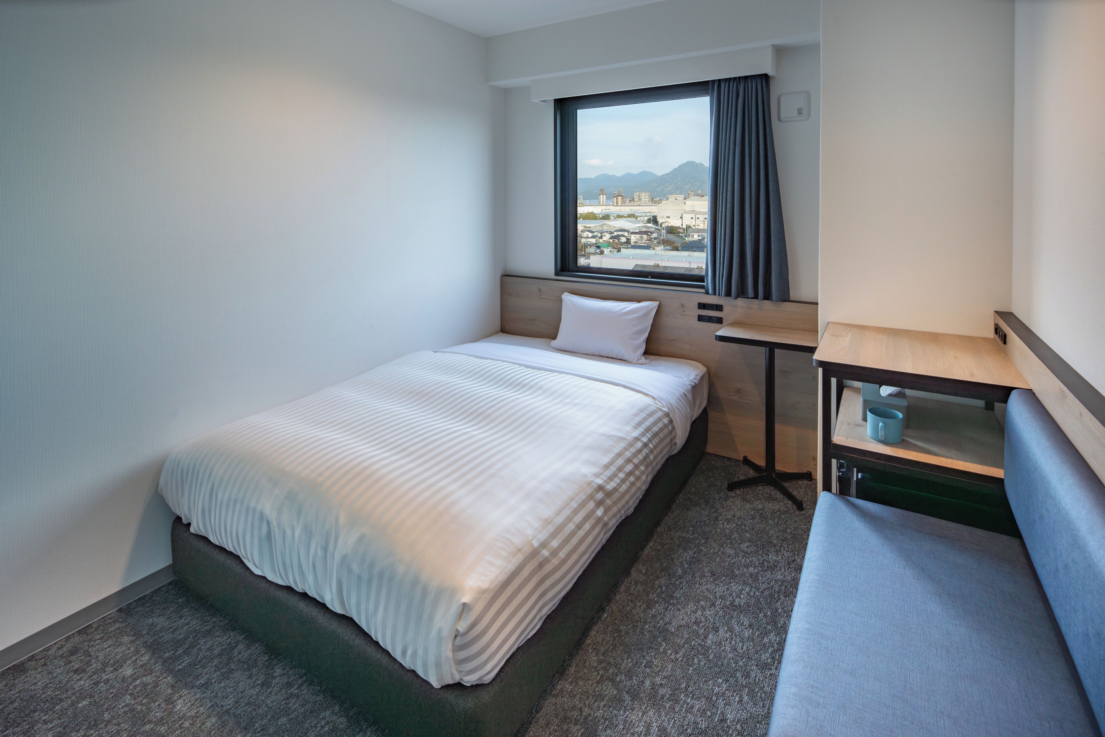 Habitación de hotel con cama, una mesita, un banco, una ventana con vistas y un interruptor de luz en la pared.