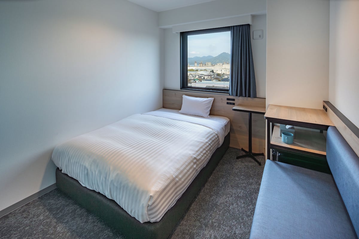 A hotel room with a bed, a small table, a bench, a window with a view, and a light switch on the wall.