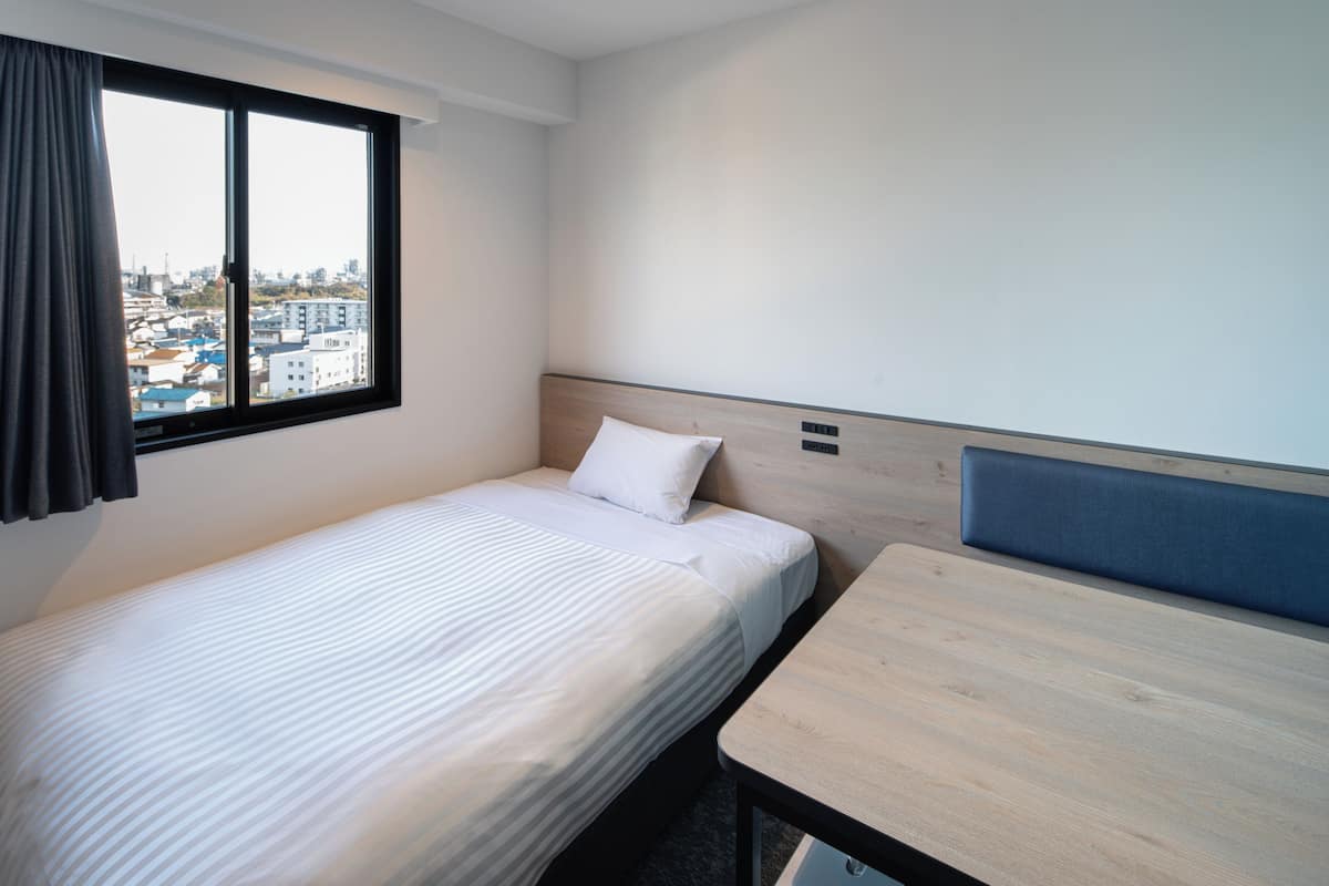 A hotel room with a single bed, a small table, a window with curtains, and a city view.