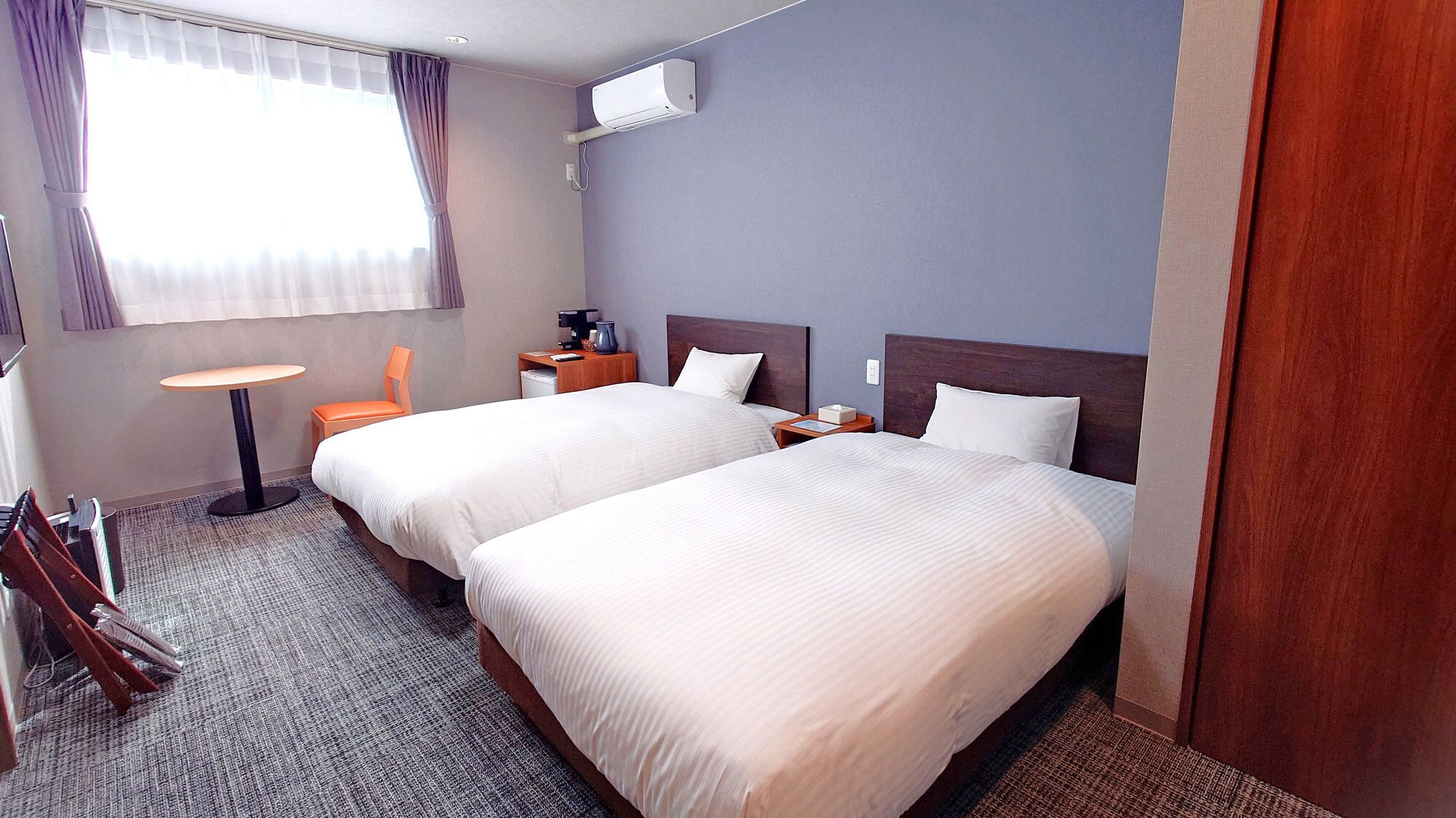 Habitación de hotel con dos camas, una mesita, una silla naranja, un escritorio y una ventana con cortinas.
