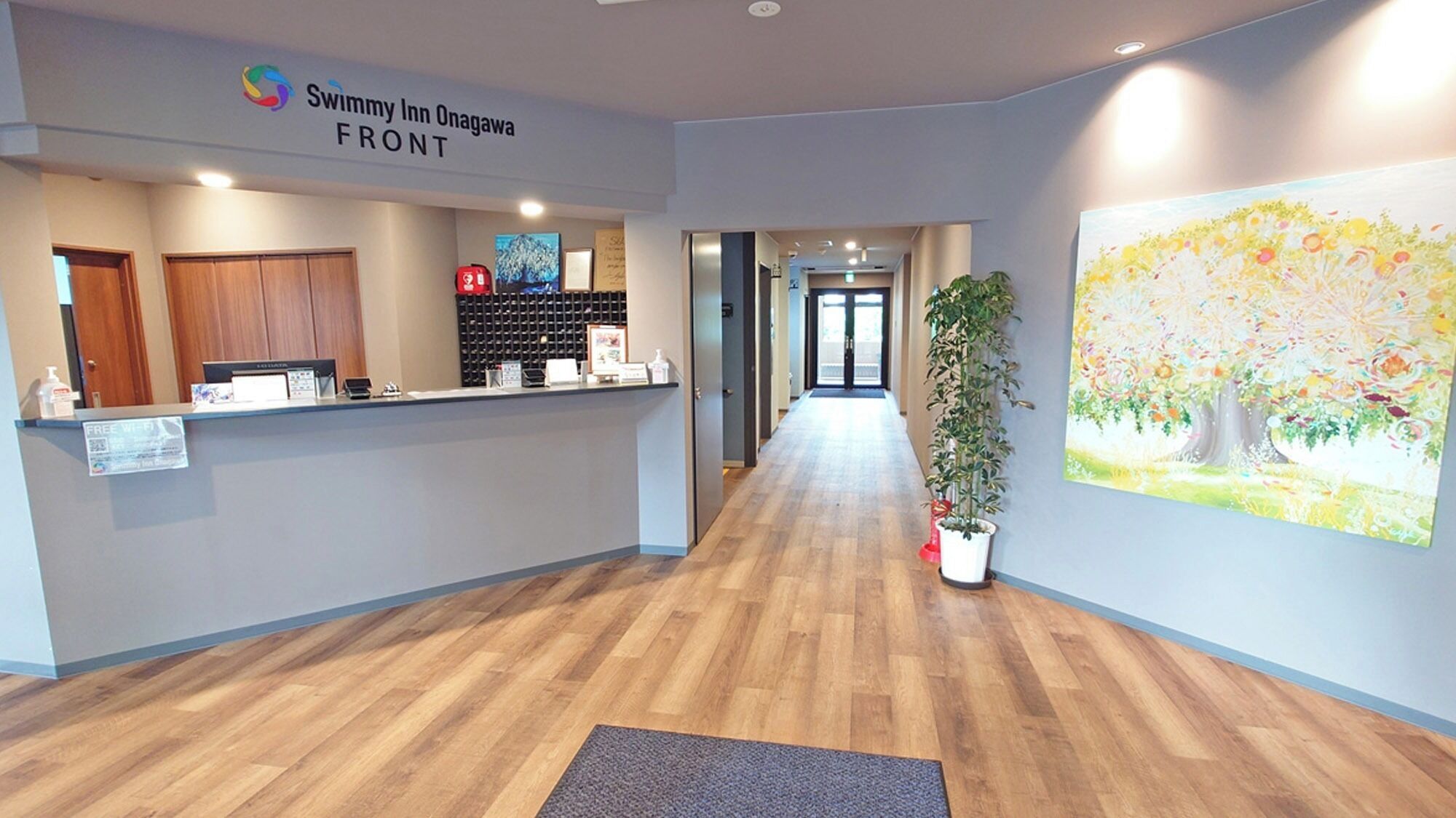 Un área de recepción con piso de madera, un mostrador de recepción, un cartel que dice 'Swimmy Inn Onagawa FRONT', una planta en maceta y un colorido mural.