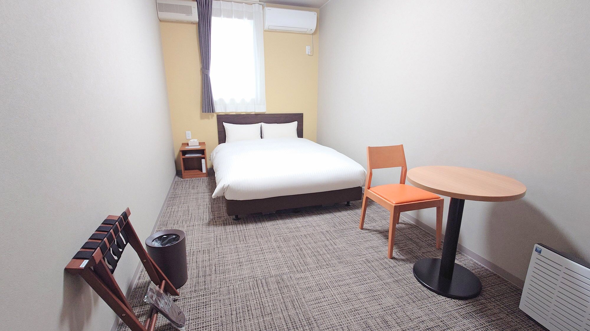 Habitación de hotel con una cama, una silla, una mesa, un cesto de basura y un equipo de aire acondicionado.
