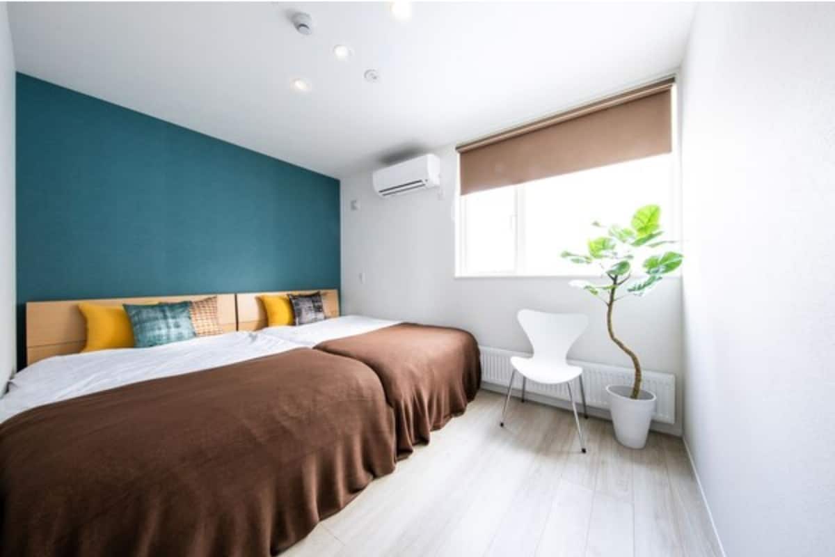 Une chambre d’hôtel moderne, équipée d’un lit, d’une chaise, d’une plante en pot et d’une fenêtre avec des stores.