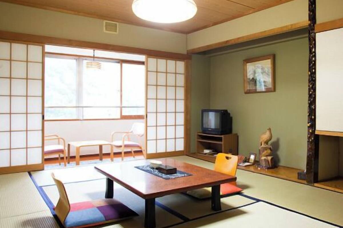 Ein Raum mit Tatami-Matten, einem Holztisch, Stühlen, einem Fernseher und einer dekorativen Skulptur.