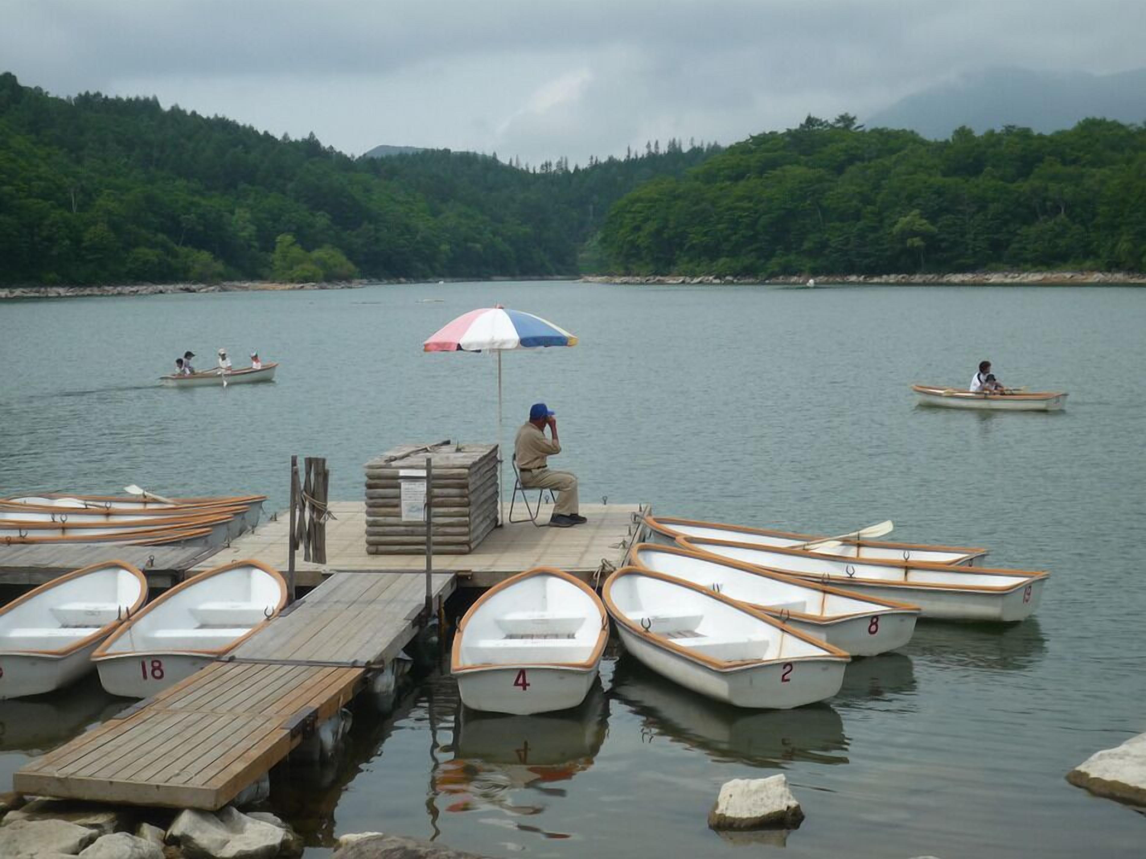 Un muelle con varios botes y un hombre sentado bajo una sombrilla, con una ladera boscosa al fondo.
