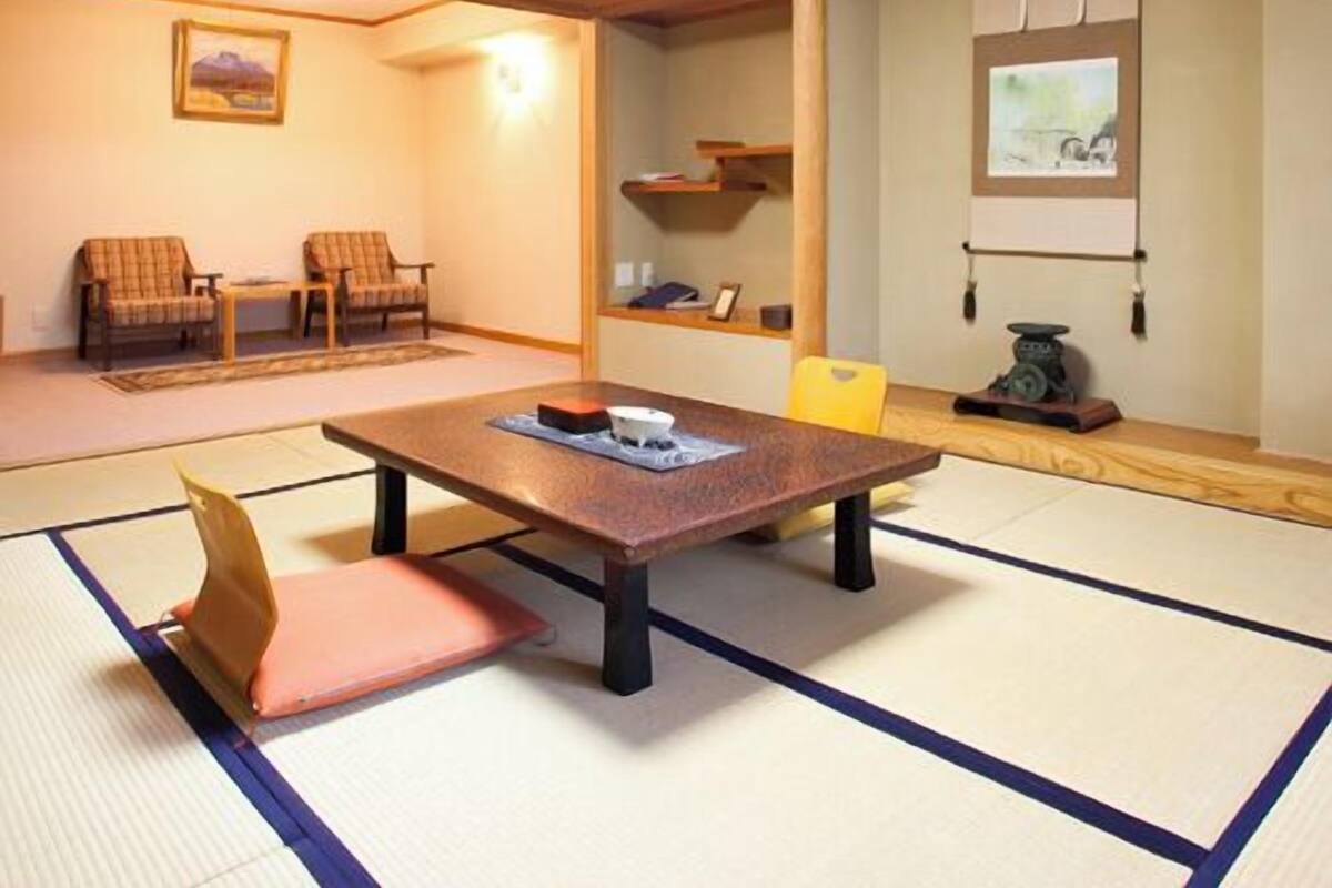 Ein Raum mit Tatami-Matten, einem niedrigen Tisch, zwei Stühlen, einem Regal mit Gegenständen und einem Fenster mit Jalousien.