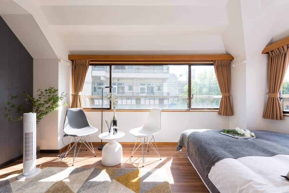 Ein modernes Hotelzimmer mit großem Fenster, einem Bett, zwei weißen Stühlen, einem kleinen Tisch mit einer Vase und einer Topfpflanze.