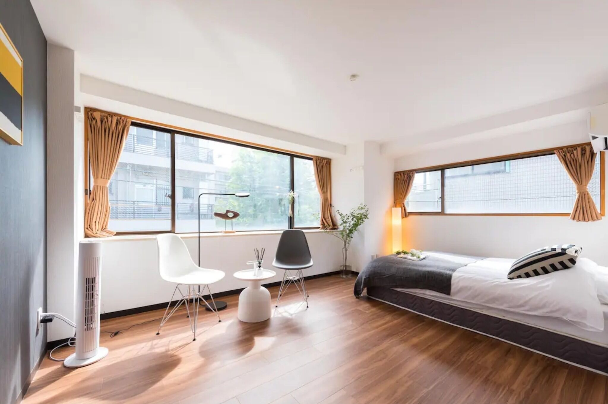 Una habitación de hotel moderna con una cama, un escritorio, dos sillas, una lámpara de pie y ventanas grandes con cortinas.