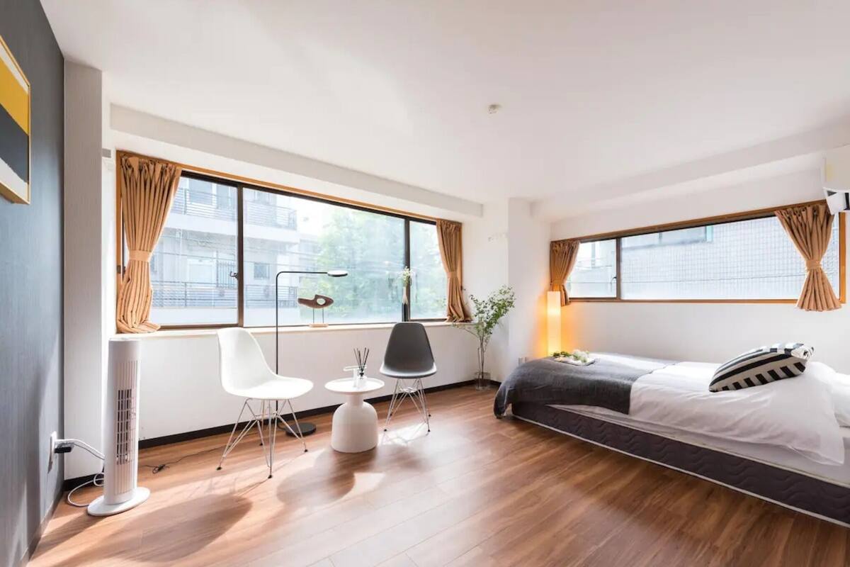 Ein modernes Hotelzimmer mit einem Bett, einem Schreibtisch, zwei Sesseln, einer Stehlampe und großen Fenstern mit Vorhängen.