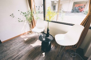 A room with two white chairs, a small round table, a vase with yellow flowers, and a view of the street outside. - Tora Hotel Rikugien (Tokyo)