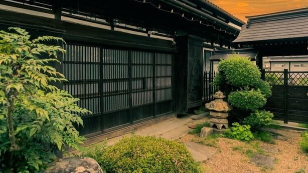 A traditional Japanese building with a black gate, a stone lantern, and a small garden with bamboo and shrubs. - Maruki Saifuku (Kakuda)