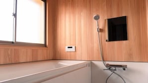 A modern bathroom with a wooden wall, a built-in bathtub, a showerhead, and a control panel. - Maruki Saifuku (Kakuda)