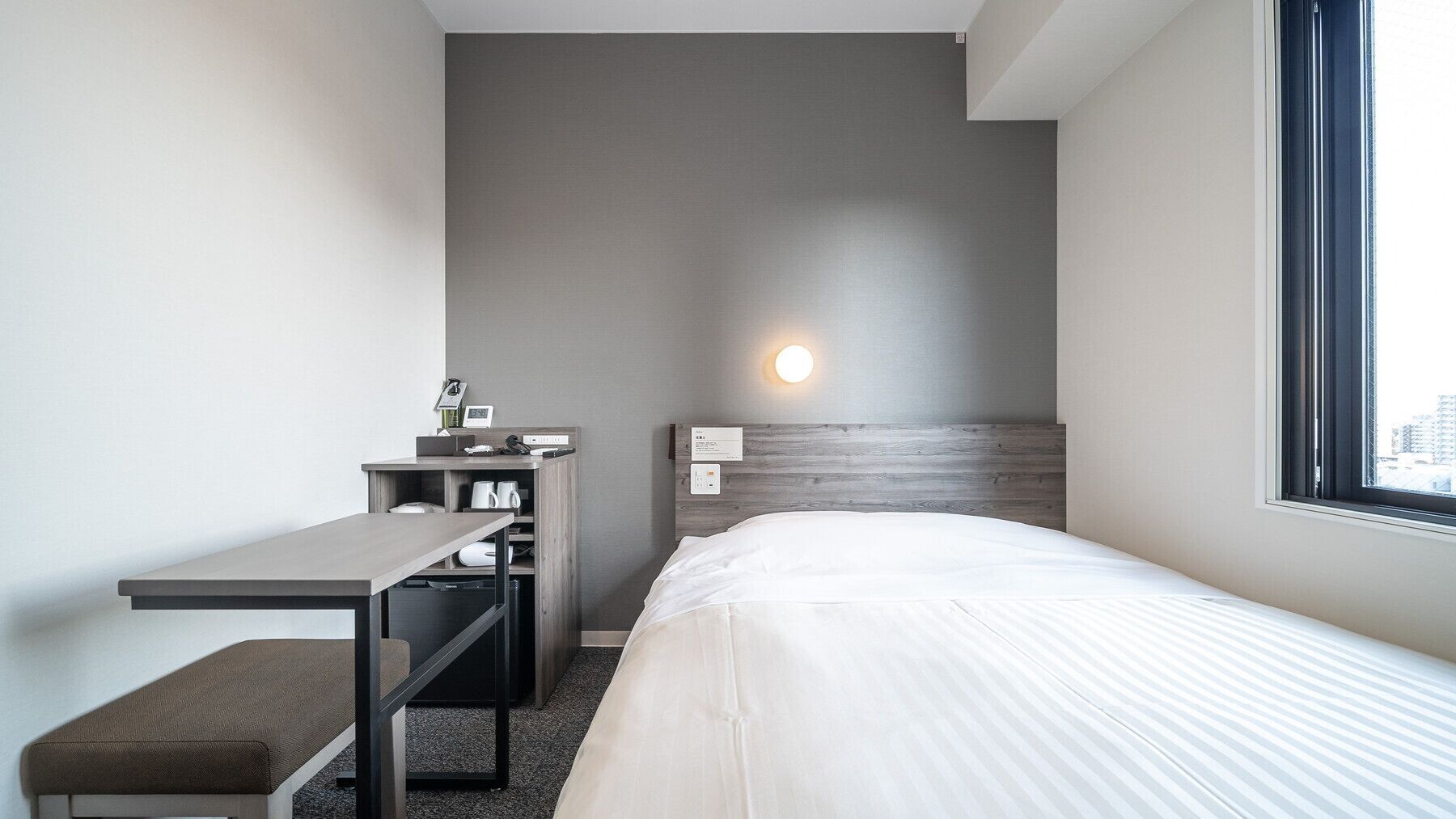 Une chambre d’hôtel comprenant un lit, un bureau, une chaise, une table de chevet, une lampe fixée au mur et une fenêtre.