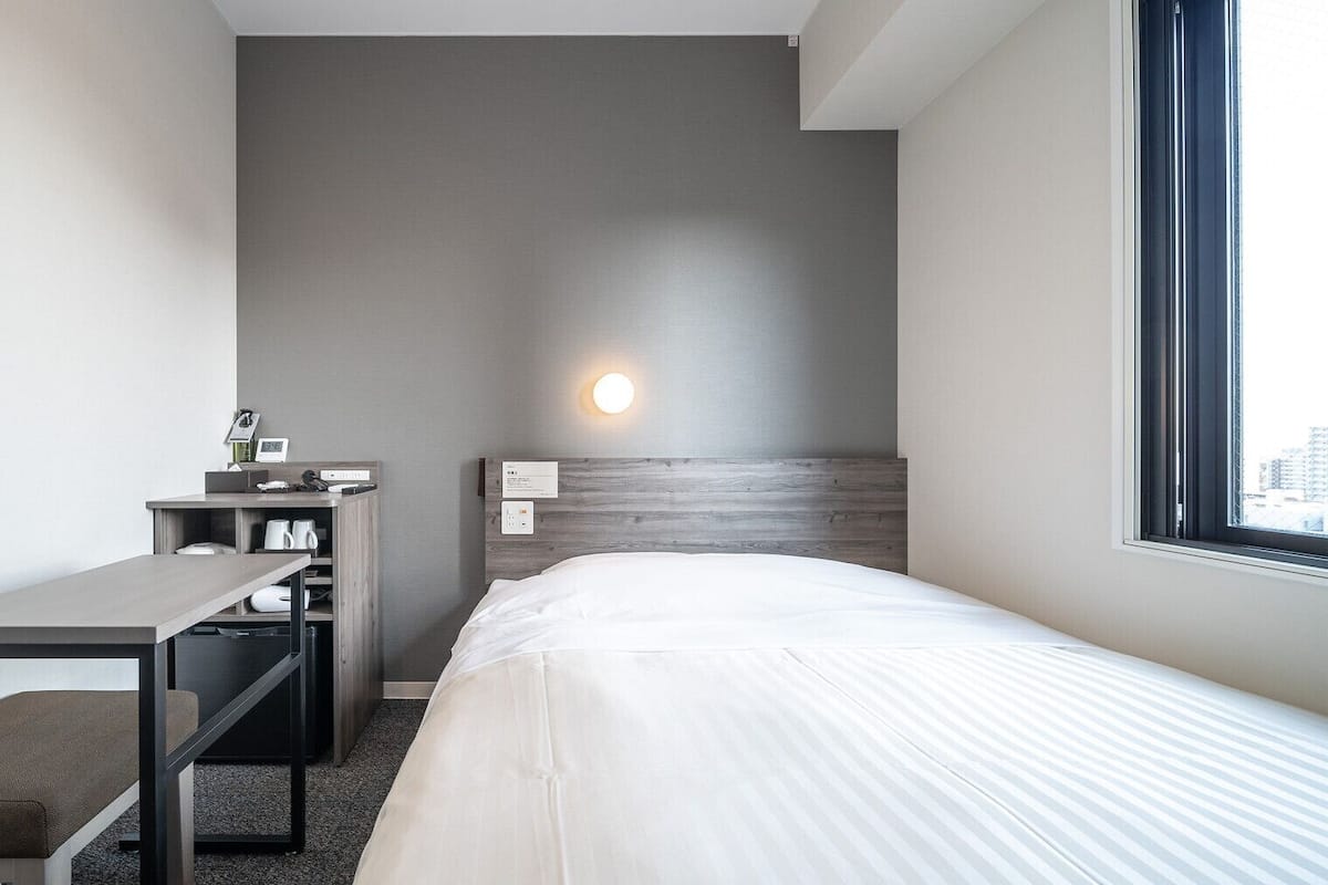 Hotellihuone, jossa on sänky, työpöytä, tuoli, yöpöytä, seinään kiinnitetty valaisin ja ikkuna.
