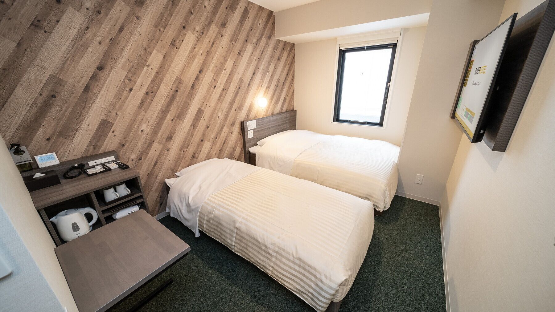 Une petite chambre d’hôtel avec deux lits, un bureau avec une chaise, une bouilloire et une fenêtre.