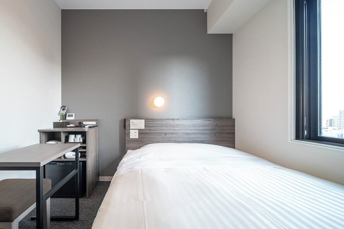 A hotel room with a bed, a desk, a chair, a nightstand, and a window.