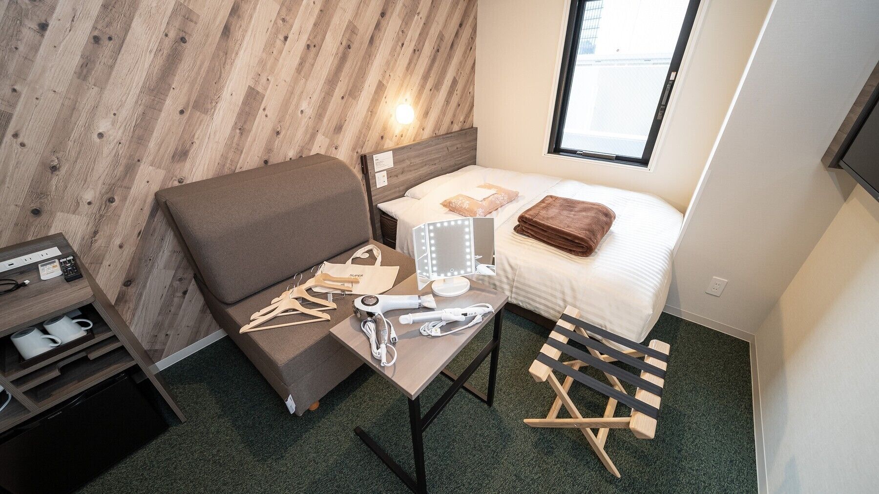Une chambre d’hôtel compacte avec un mur en bois, un lit recouvert d’une couette blanche, un canapé marron, une coiffeuse avec un miroir et une petite table sur laquelle sont posés des effets personnels.