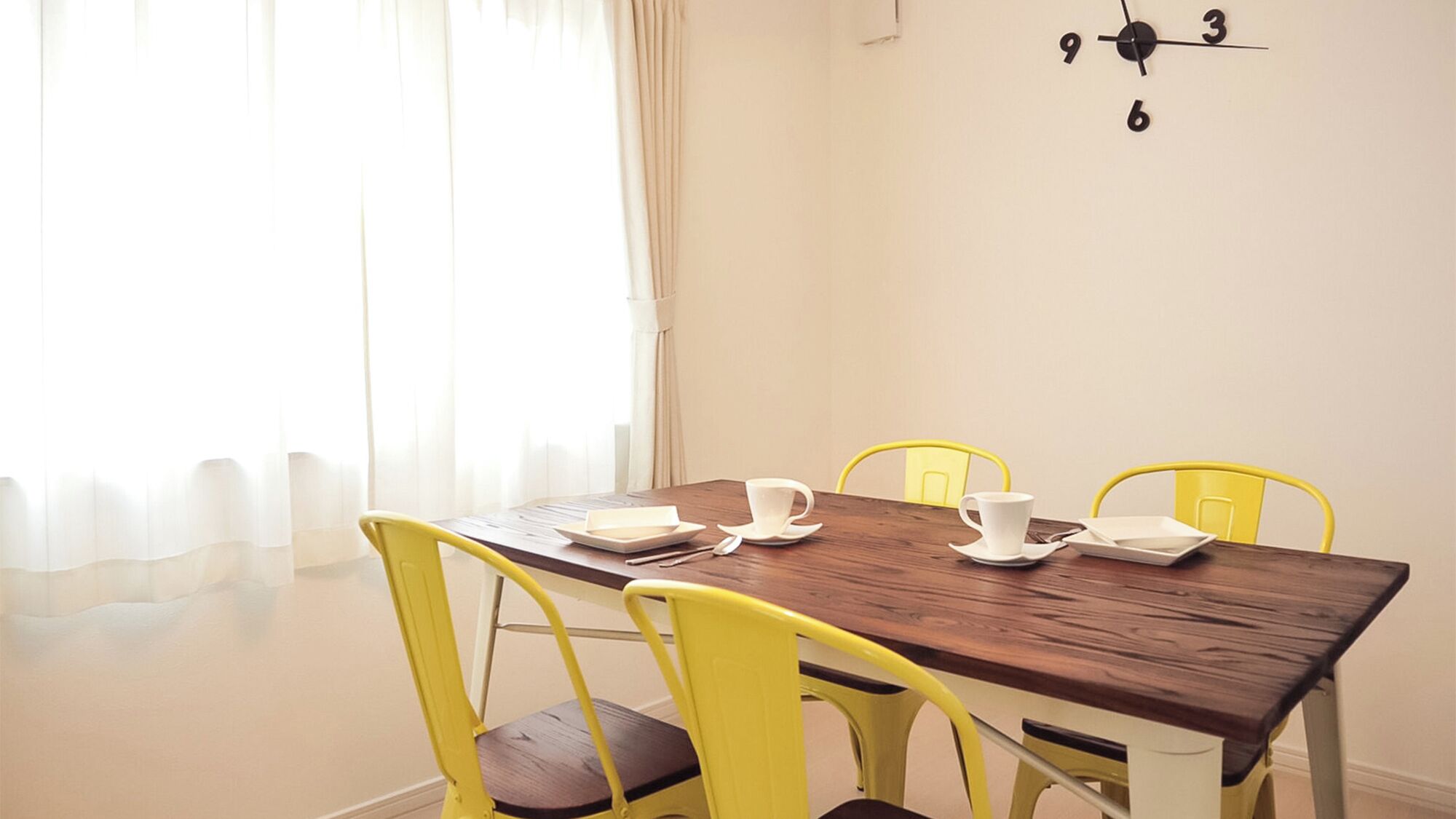 A dining table with four yellow chairs, white plates, and cups, a clock on the wall, and sheer curtains.