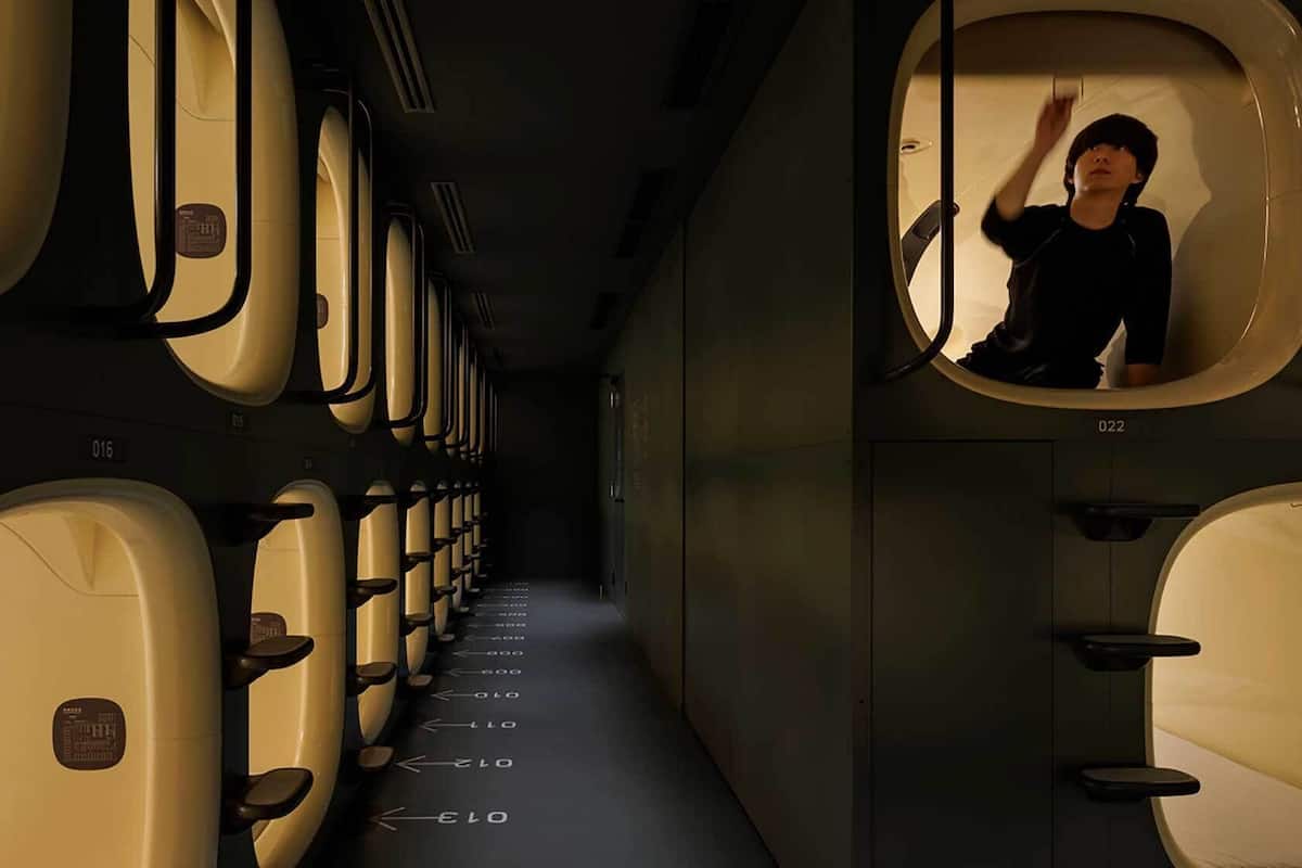 Una fila de baños de avión con puertas numeradas, una persona parada en uno de ellos, y un pasillo que conduce al fondo del avión.