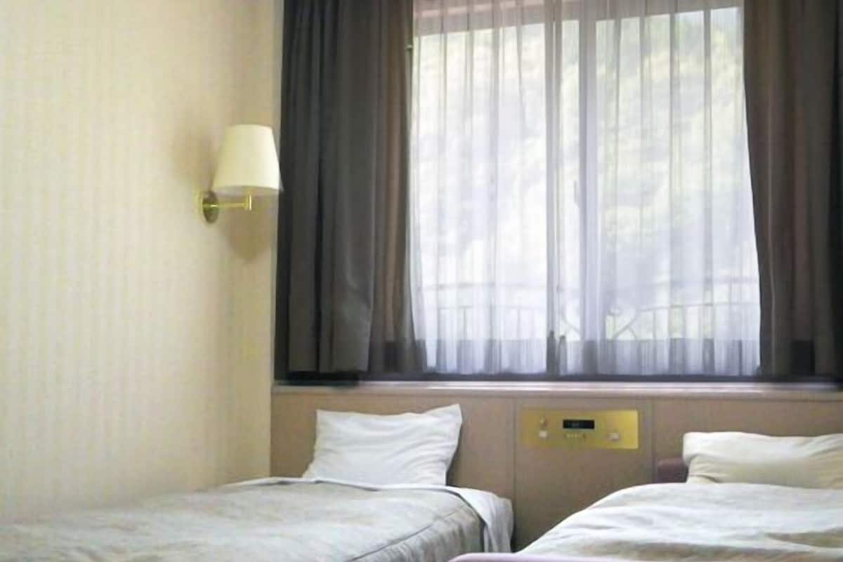 Et hotelværelse med to senge, et vindue med gardiner, en vægmonteret lampe og et kontrolpanel på væggen.