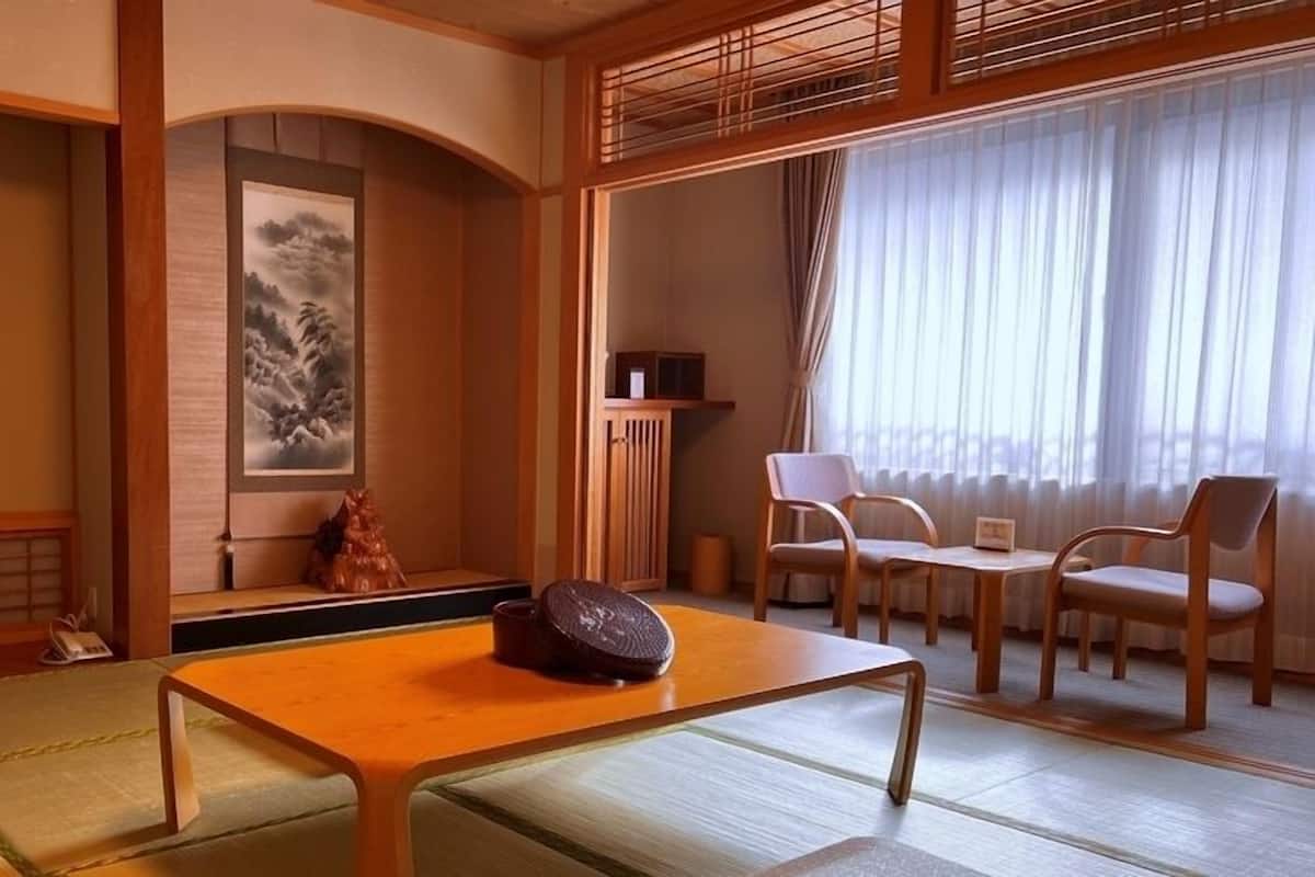 Et traditionelt japansk rum med tatamimåtter, et lavt bord med en dekorativ genstand, et maleri og udsigt gennem lette gardiner.