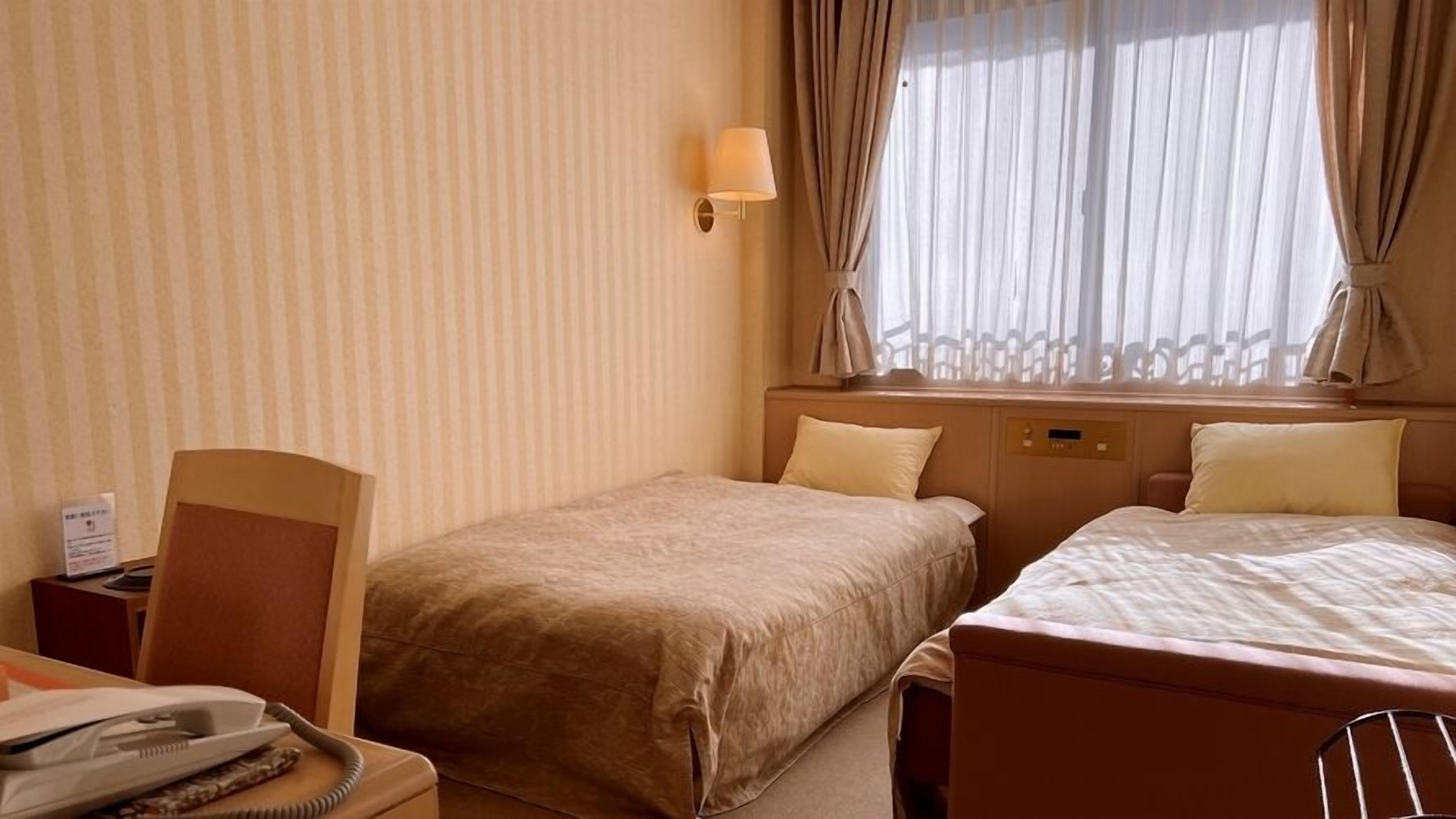 A hotel room with two beds, a bedside table with a lamp, a telephone, and a window with curtains.