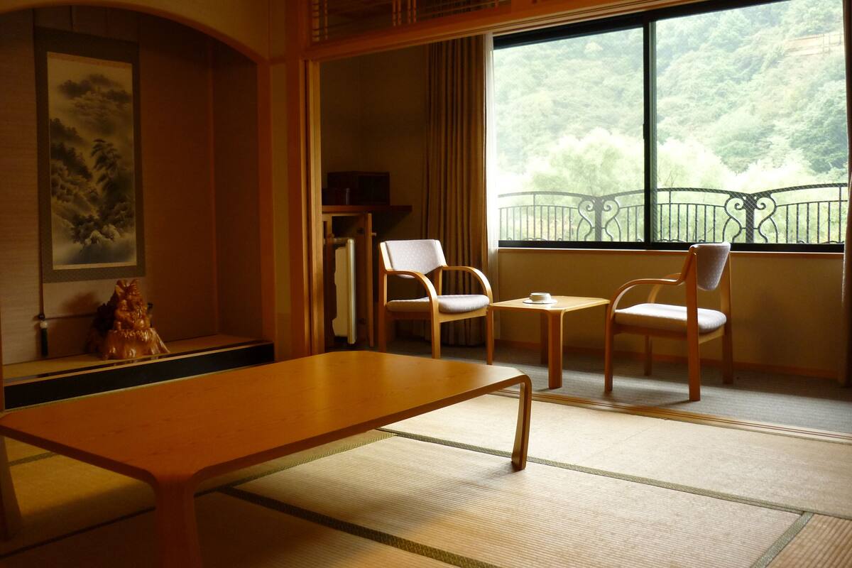 Et traditionelt japansk rum med tatamimåtter, et lavt bord, to stole, et lille bord med en kop, og udsigt til en skov fra en balkon.