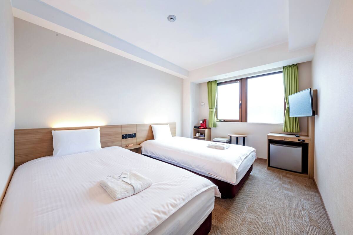 A hotel room with two beds, a small desk, a television, and a window with curtains.