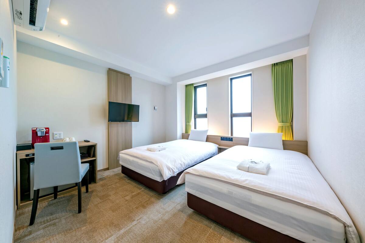 A hotel room with two beds, a desk with a chair, a TV, and a window with curtains.