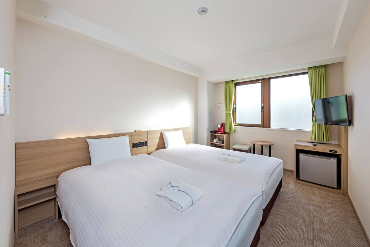 A hotel room with two beds, a television, a small desk, and a window with curtains.