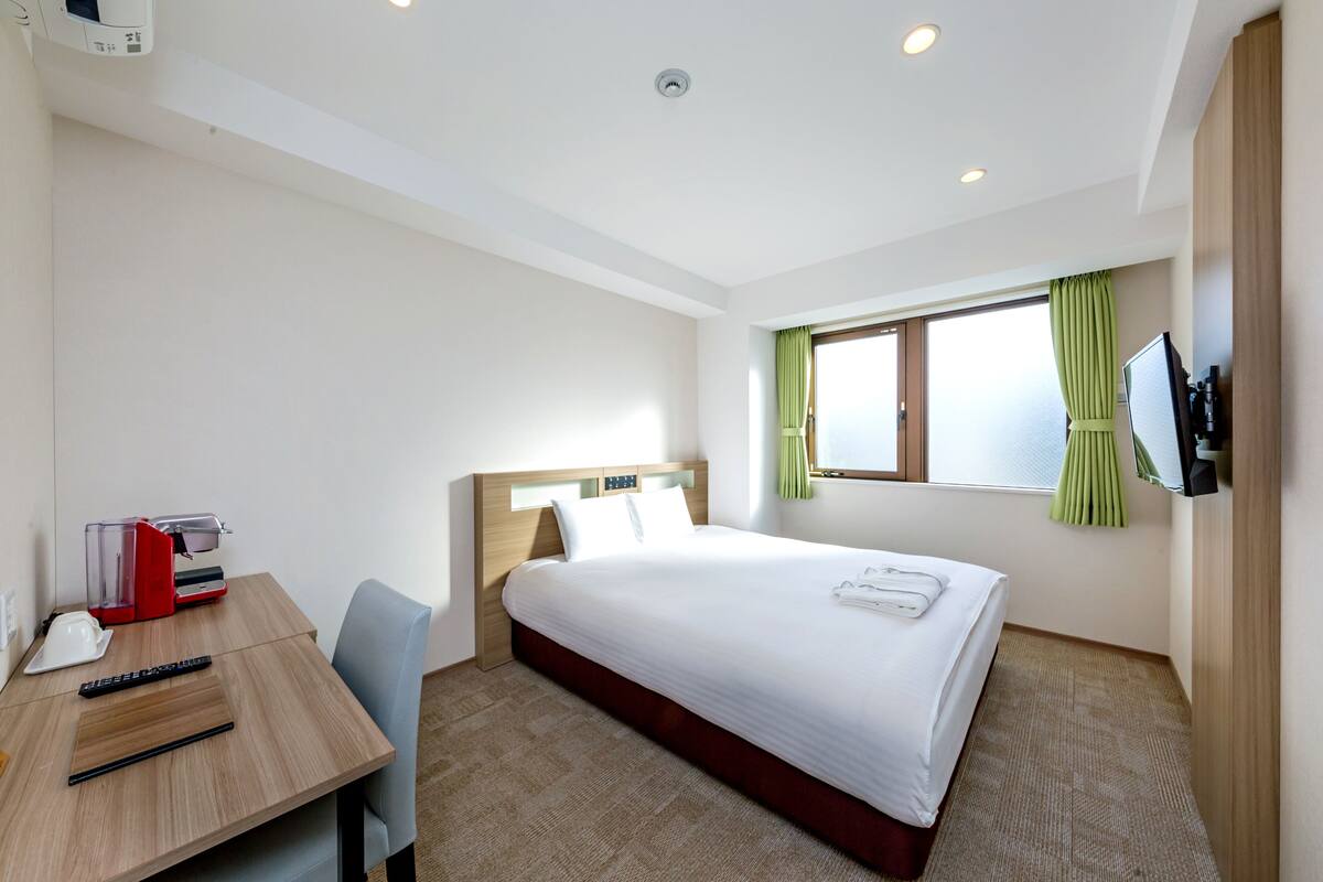 A hotel room with a bed, a desk with a computer, a TV, and a window with curtains.