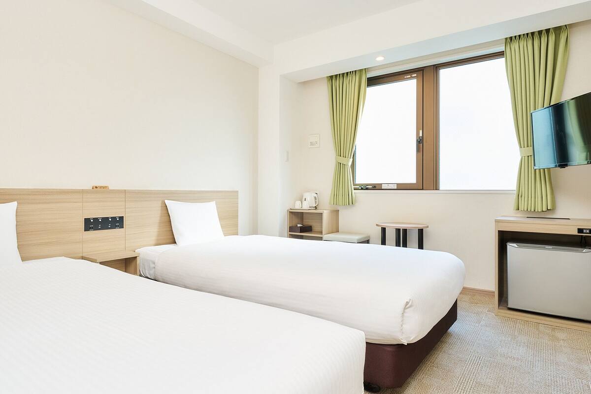 A hotel room with two beds, a small desk, a television, and a window with curtains.