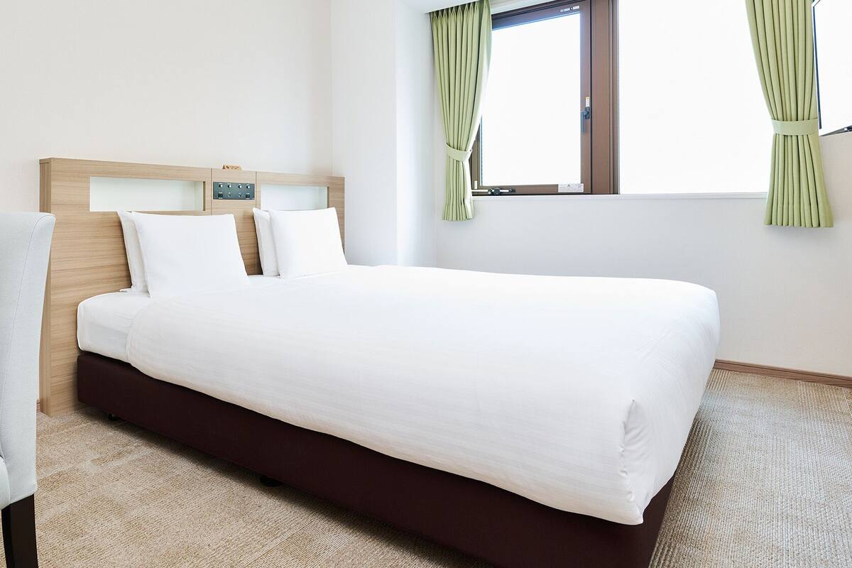 A hotel room with a bed, a chair, a window with curtains, and a wooden headboard.