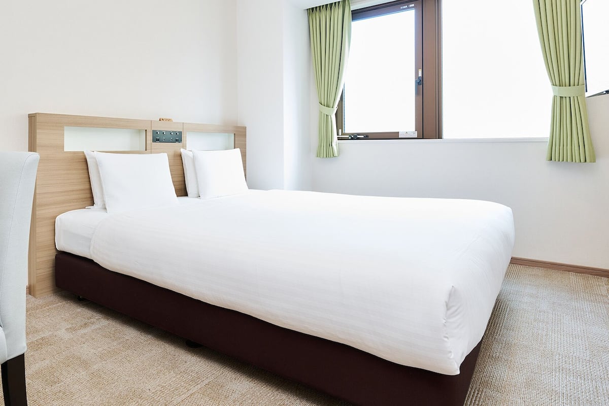 Hotellihuone, jossa on sänky, tuoli, ikkuna verhoilla ja puinen pääty.