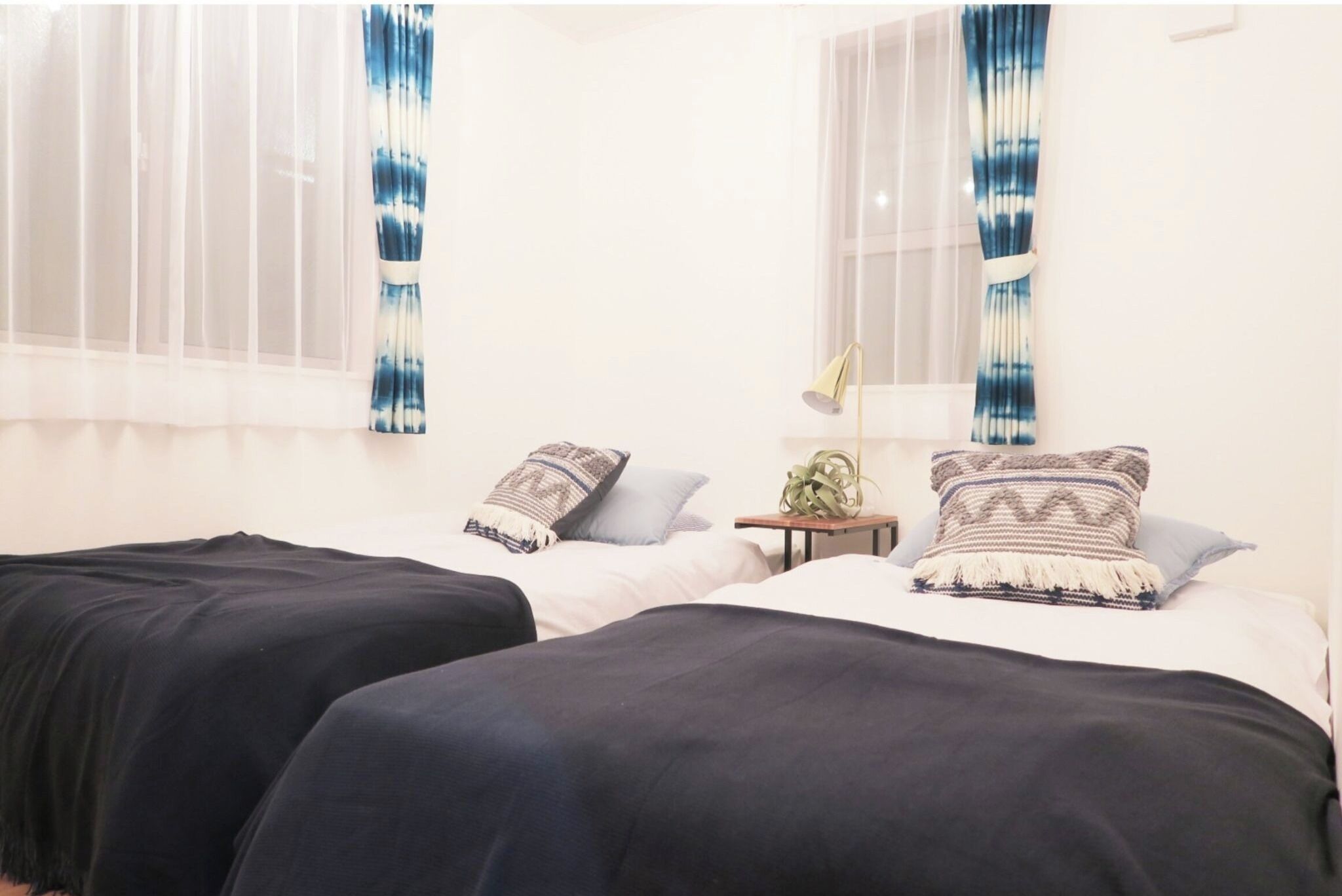 A room with two beds, white bedding, a small table with a lamp, and sheer curtains with a blue pattern.