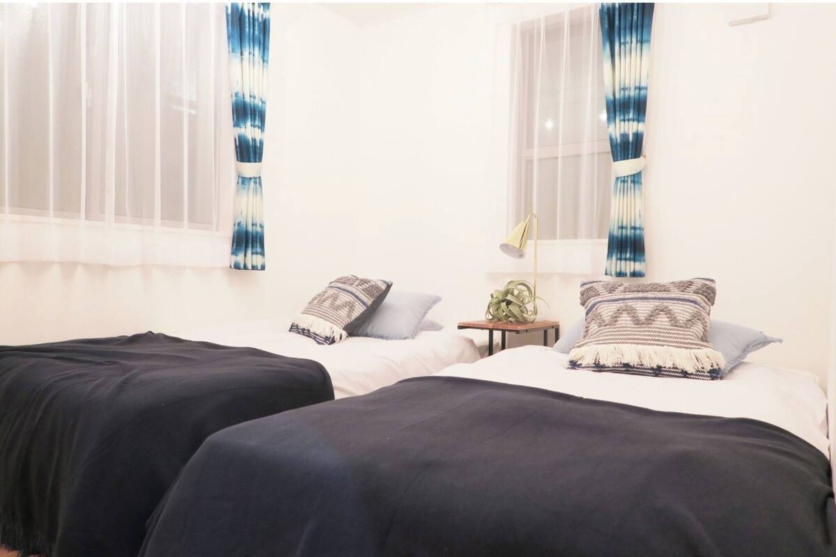 A room with two beds, white bedding, a small table with a lamp, and sheer curtains with a blue pattern.