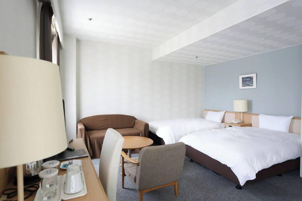 A hotel room with two beds, a desk with a lamp, a chair, and a small table.
